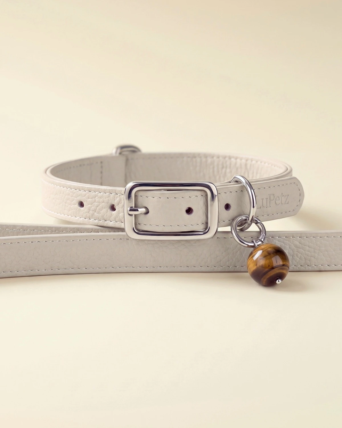 A taupe leather LuKra collar beside its matching taupe leash, which has a tiger's eye bead near the clip.