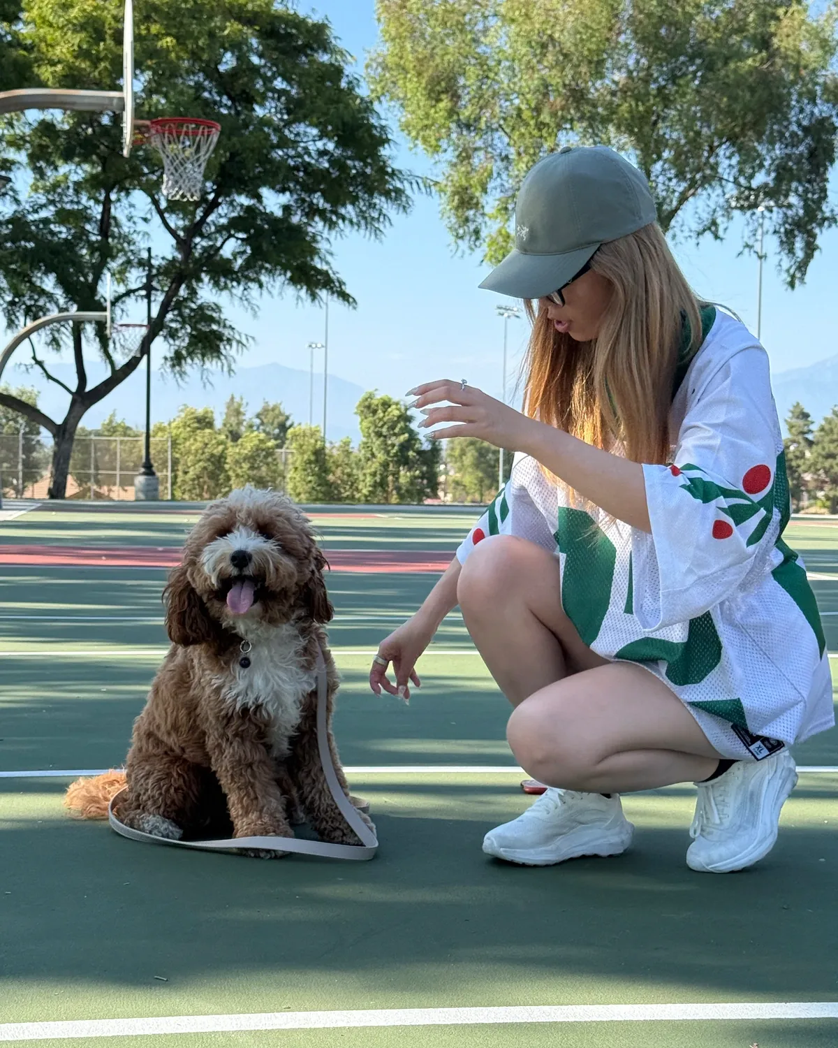 Woman in a cap kneeling beside a brown doodle on an outdoor court